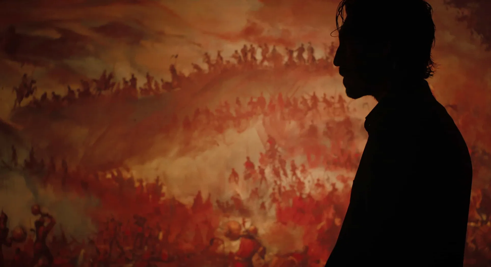 A still from Monkey Man (2024), with the main character standing in silhouette against a painting of a huge battle.